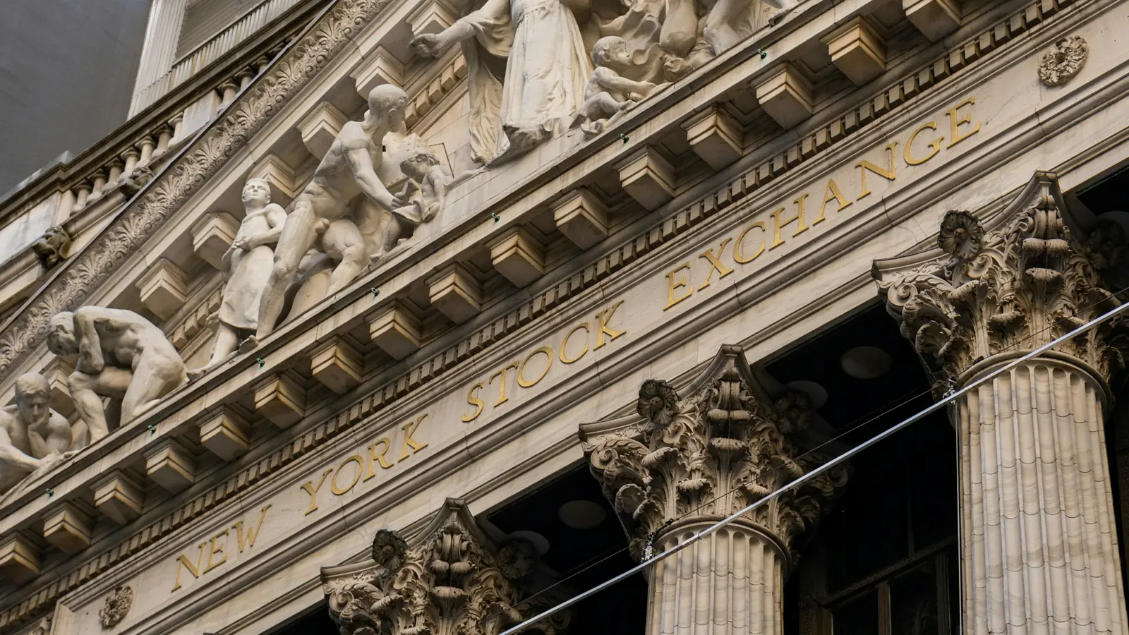 The carved pediment and Corinthian columns of the New York Stock Exchange on Broad Street, with "New York Stock Exchange" in gold letters on the facade.