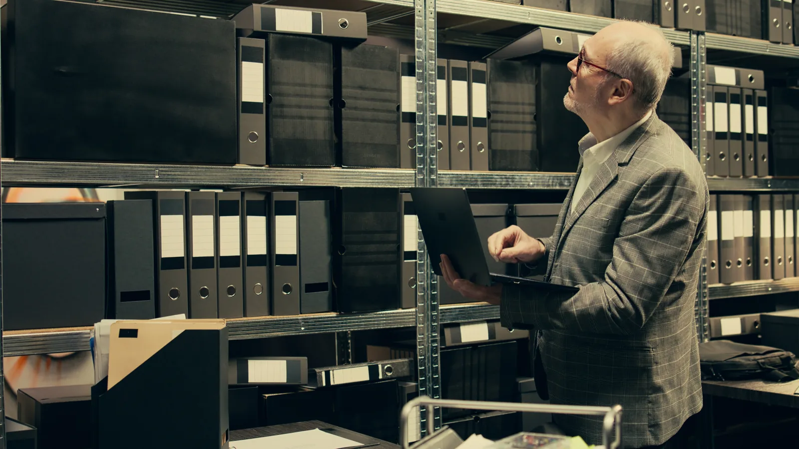A man in a gray blazer holds a laptop while scanning rows of black binders on archive shelves.